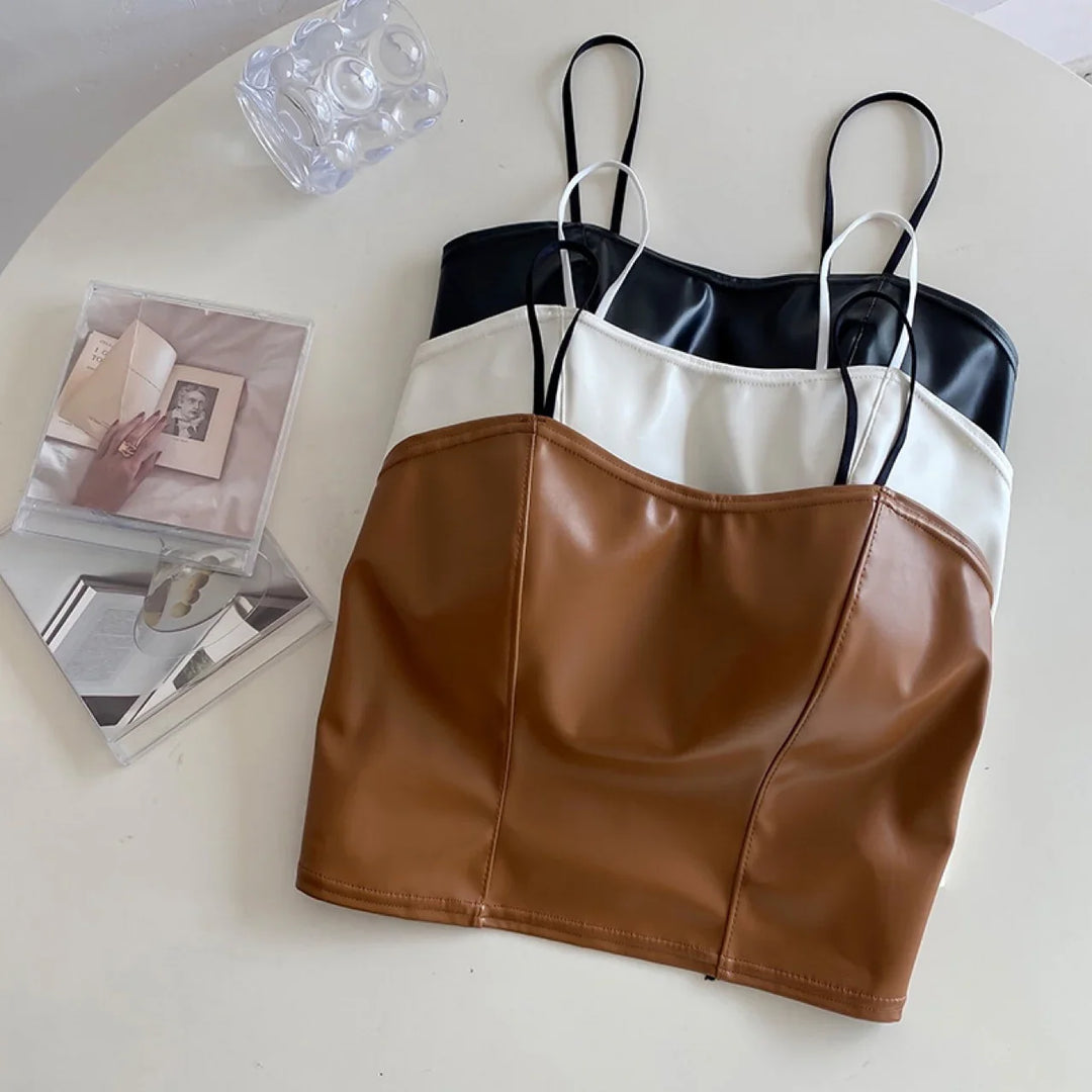 Brown, white, and black faux leather camisole tops with thin straps on a white table with glass decor and a photo book