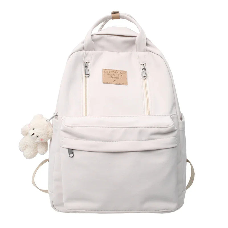 White backpack with front pockets, two zippers, beige logo patch, and small white teddy bear keychain