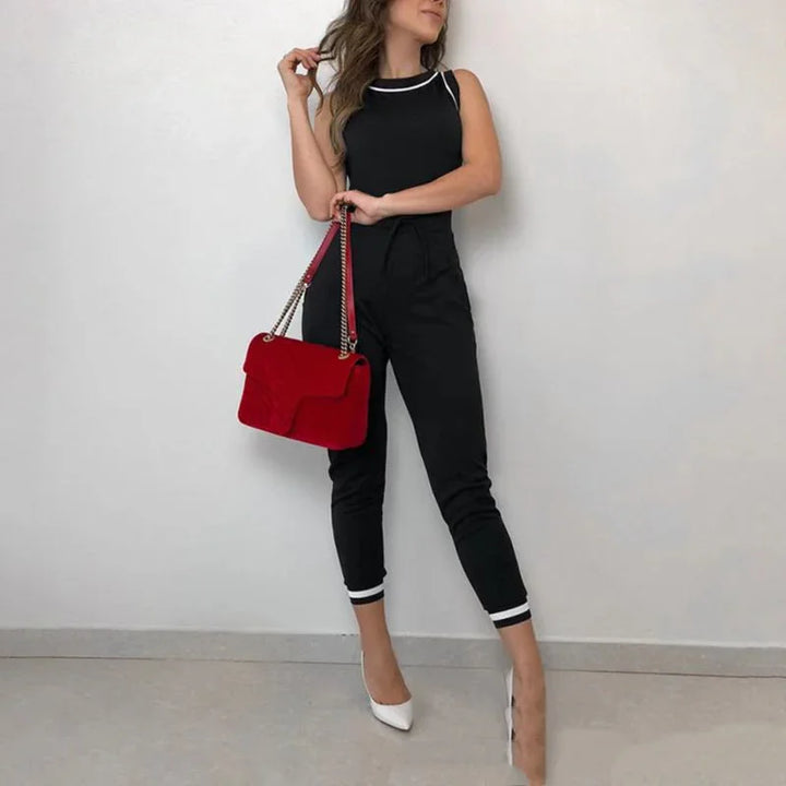 Woman in black sleeveless jumpsuit with white trim holding red shoulder bag and wearing white heels