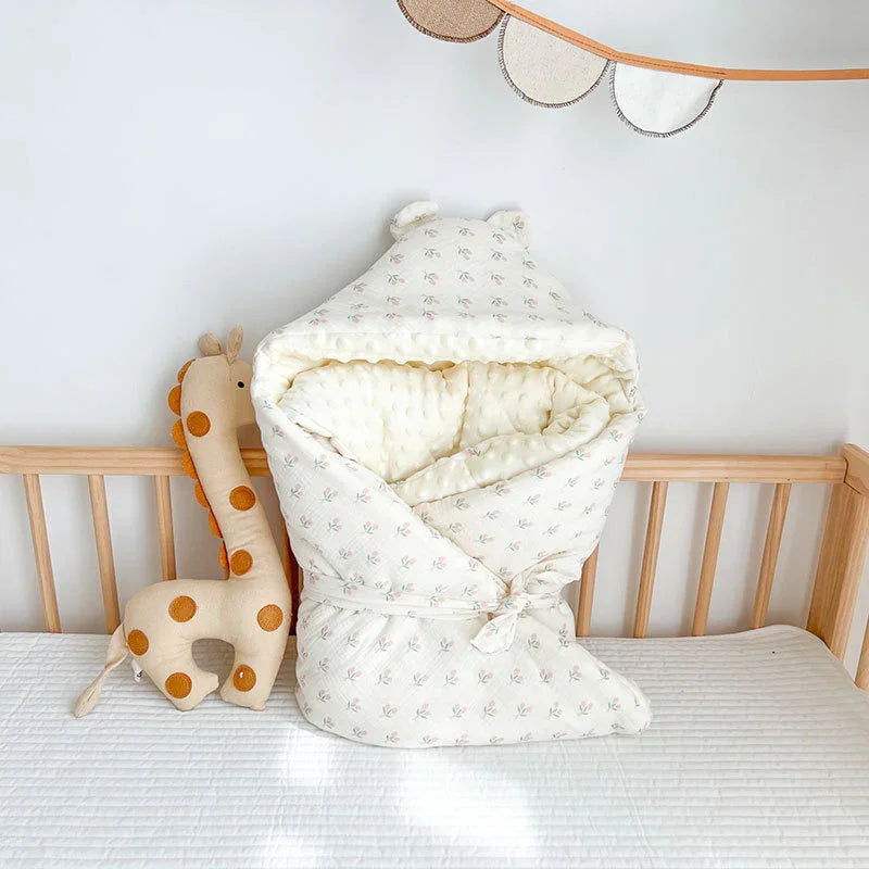 Cozy baby wrap blanket with ears and floral print on crib mattress next to plush giraffe toy in nursery