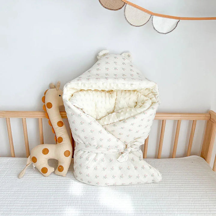 Cozy baby wrap blanket with ears and floral print on crib mattress next to plush giraffe toy in nursery