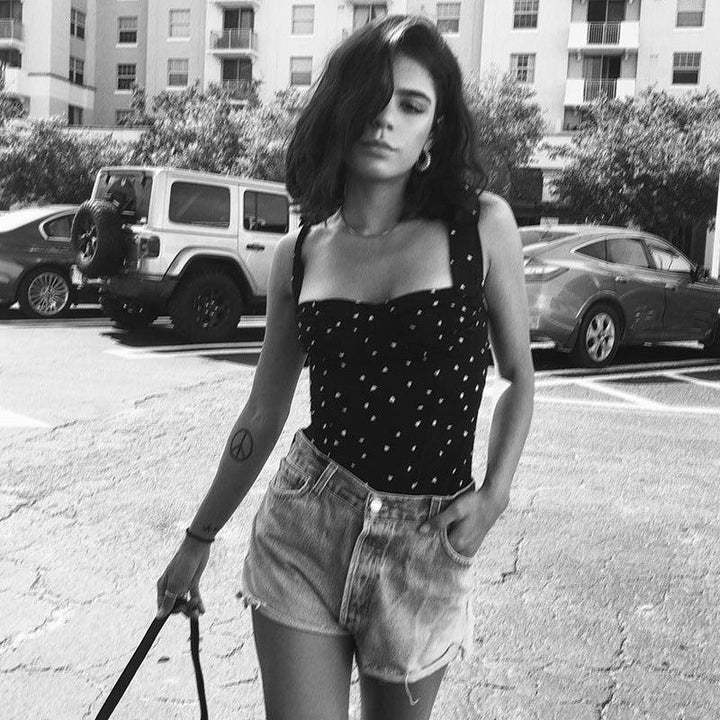 Young woman in patterned black top and denim shorts posing in urban parking lot with cars and apartment buildings