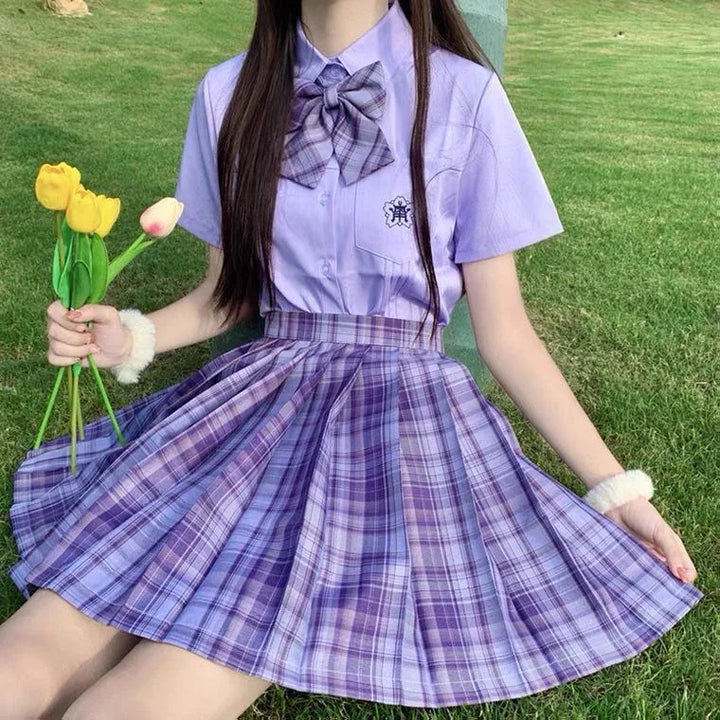 Woman wearing purple plaid pleated skirt and matching bow tie shirt sitting on grass holding yellow tulips