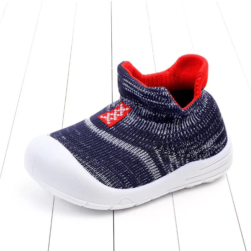 Navy blue knitted baby shoe with white sole and red interior lining on white wooden floor