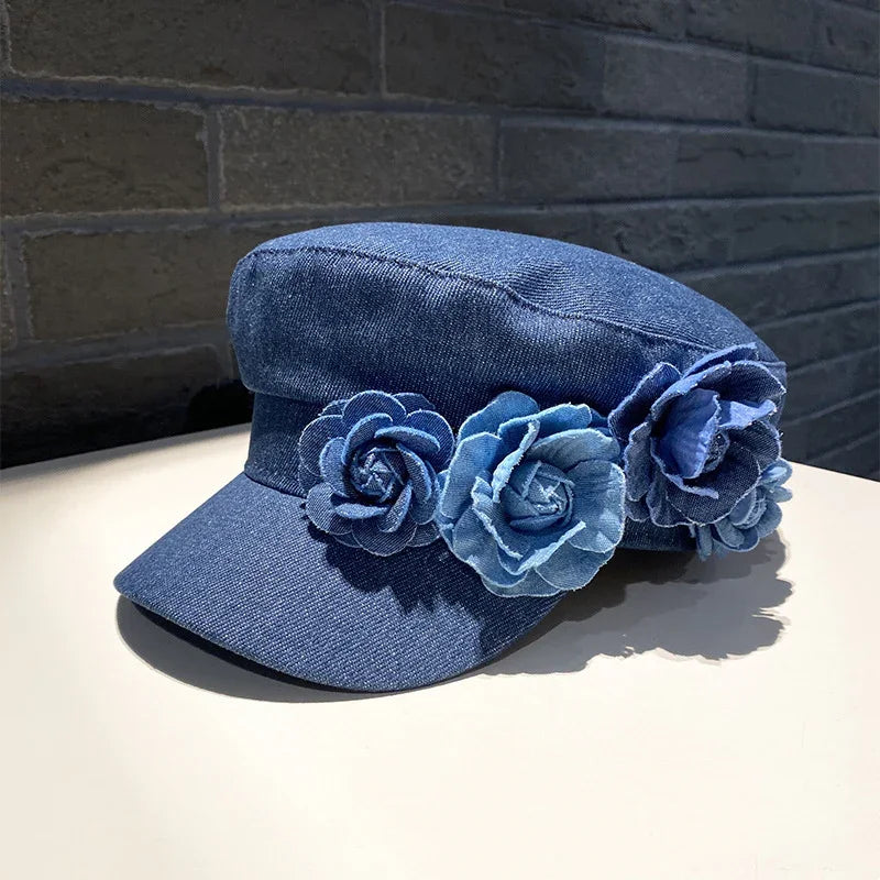 Blue denim cap with three large denim flower appliques on the side against a dark brick background