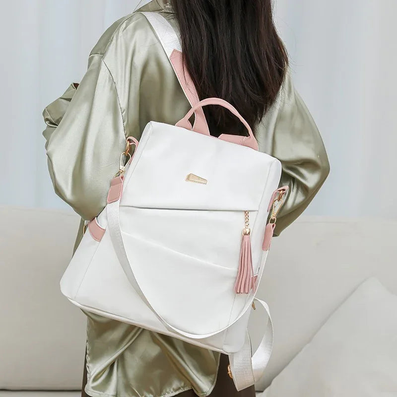 White and pink backpack with tassel detail worn by person in light green satin jacket indoors