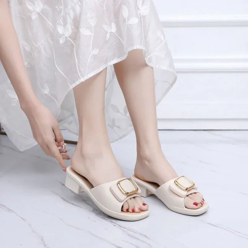 Woman wearing beige heeled slip-on sandals with gold buckle, white floral dress, on marble floor
