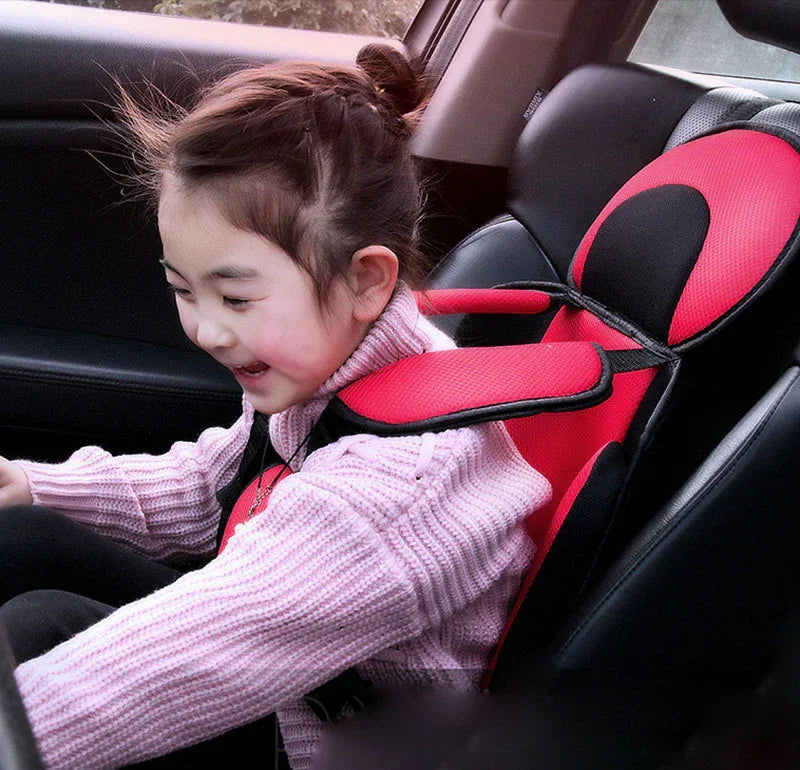 Smiling child sitting in red and black car seat with armrests, wearing pink sweater inside a car
