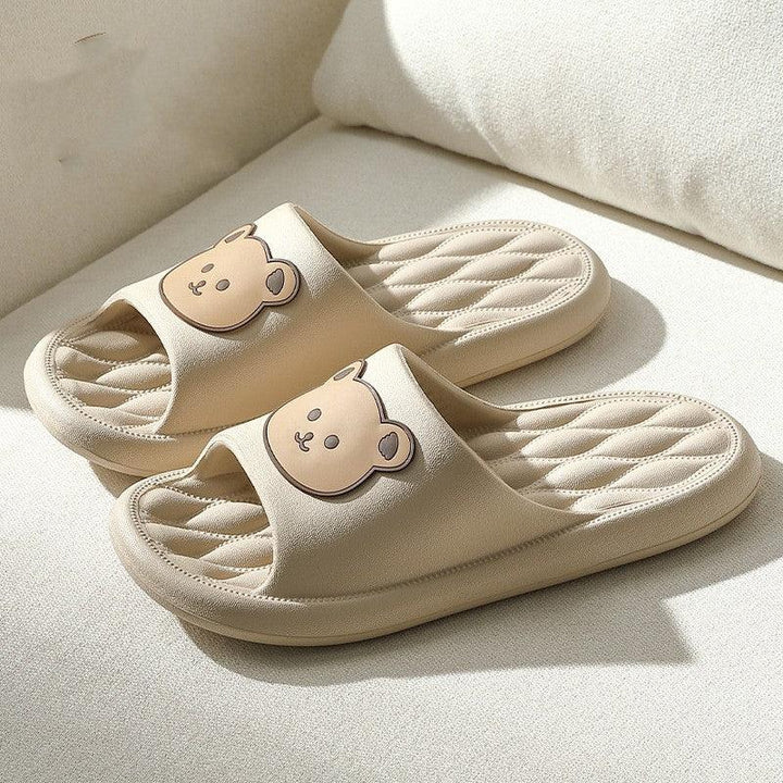 Beige cushioned slippers with cute bear face design on top, placed on white fabric surface
