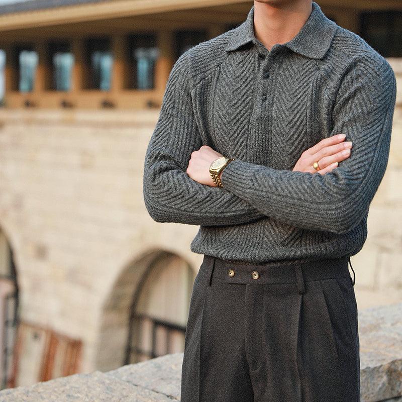 Man wearing textured grey knit sweater and dark pleated trousers with gold watch standing outdoors