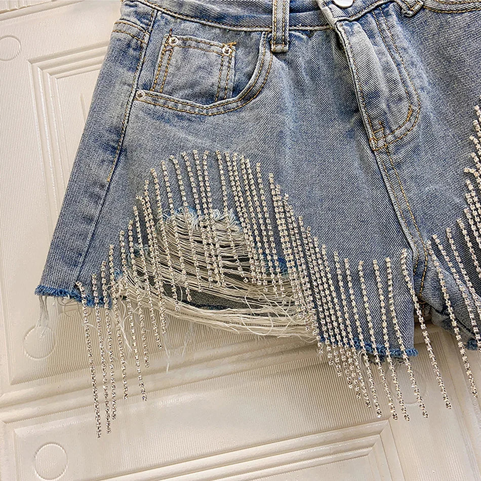 Light blue distressed denim shorts with rhinestone fringe detailing and raw hem hanging on white door