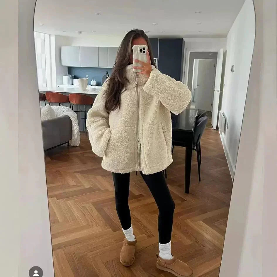 Woman wearing cream fleece jacket, black leggings, and brown slippers taking mirror selfie in modern living room