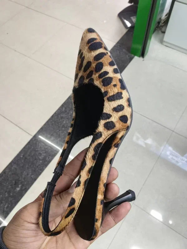 Hand holding a leopard print pointed-toe slingback high heel shoe with a black heel on tiled floor
