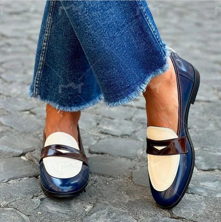 Close-up of stylish women's two-tone patent leather loafers in blue and cream with brown strap, worn with frayed hem jeans on cobblestone pavement