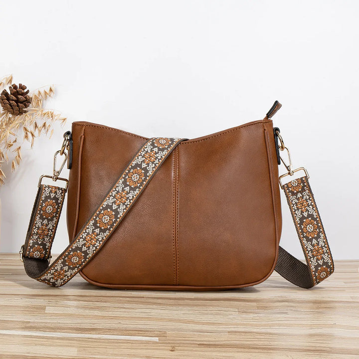 Brown leather crossbody bag with decorative floral patterned fabric strap on wooden surface