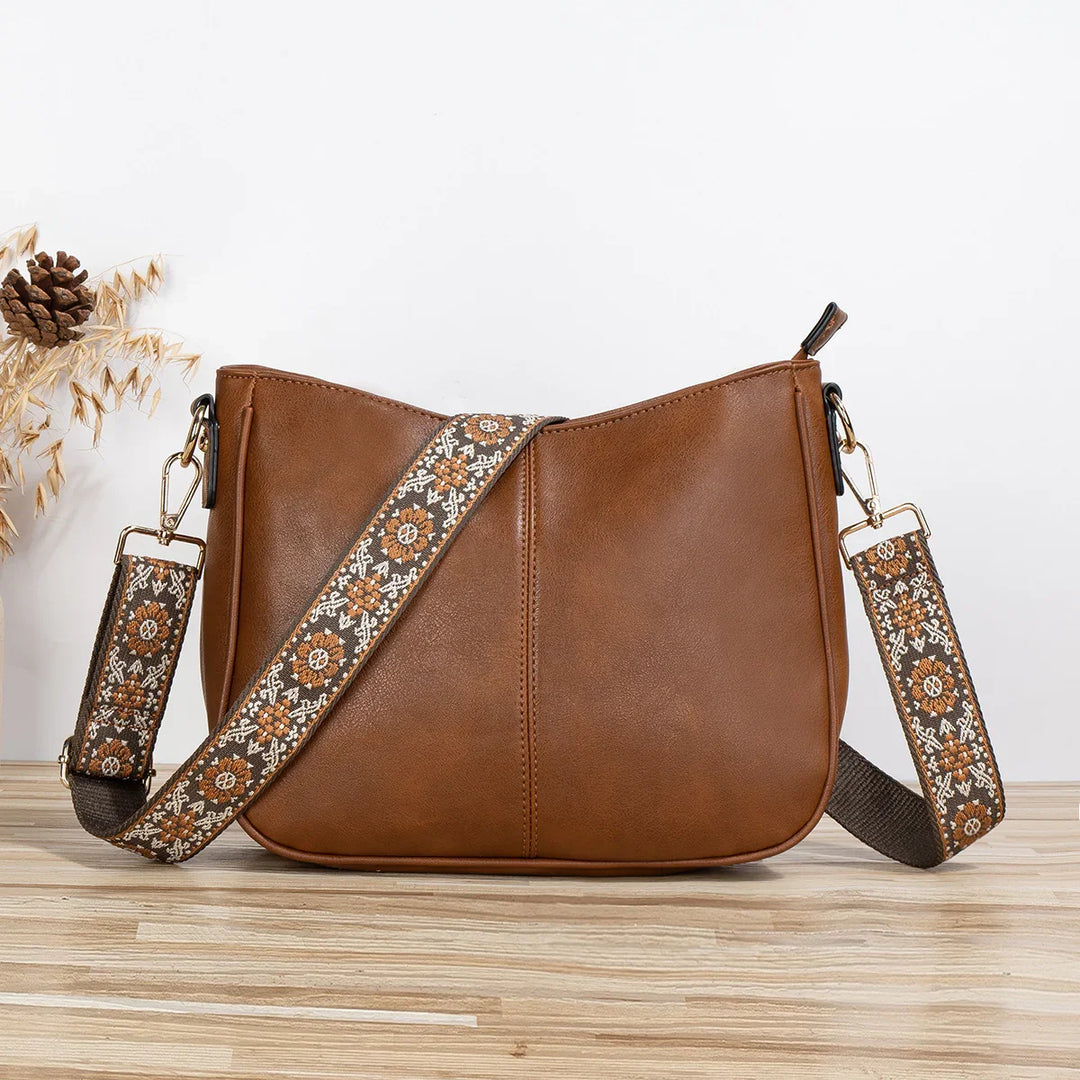 Brown leather crossbody bag with floral embroidered adjustable strap on wooden surface