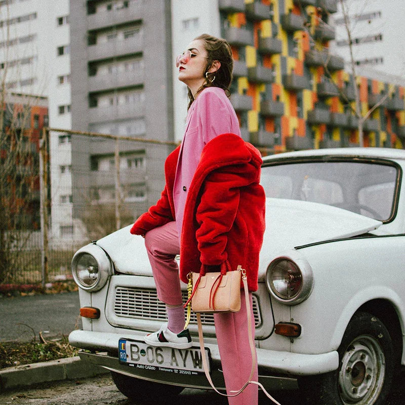 Fashionable woman in pink suit and red faux fur coat posing on vintage white car in urban setting