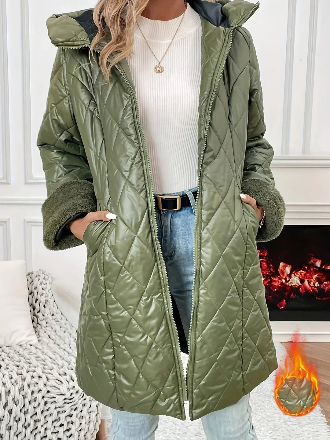 Woman wearing quilted olive green winter jacket with furry cuffs, white sweater, and jeans indoors