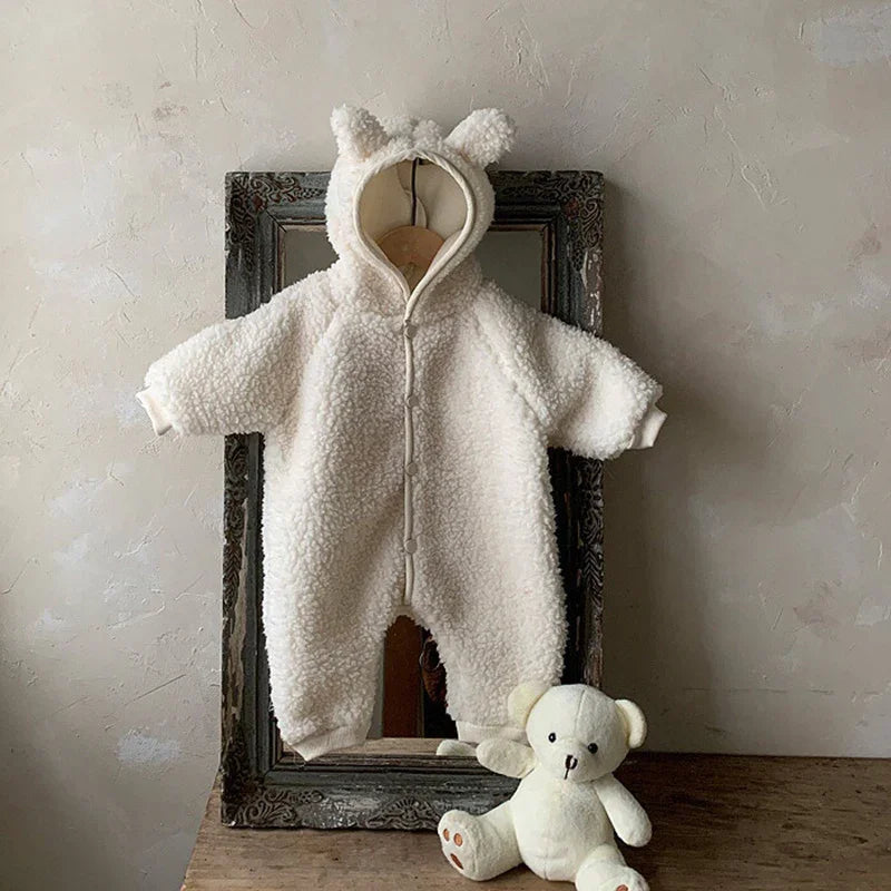 Cream-colored fuzzy baby romper with hood and ears hanging on vintage mirror frame, white teddy bear on wooden table