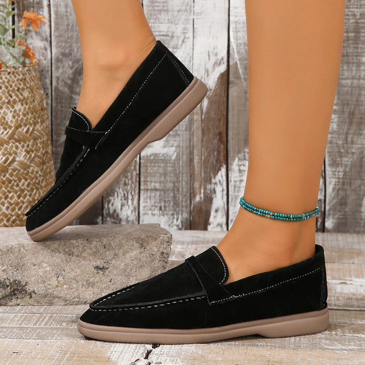 Black suede loafers with beige soles on feet, turquoise beaded anklet, rustic wooden background