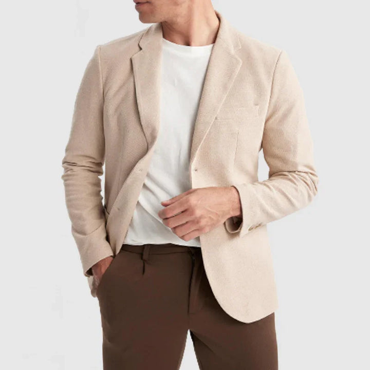 Man wearing beige blazer over white t-shirt with brown trousers on plain background