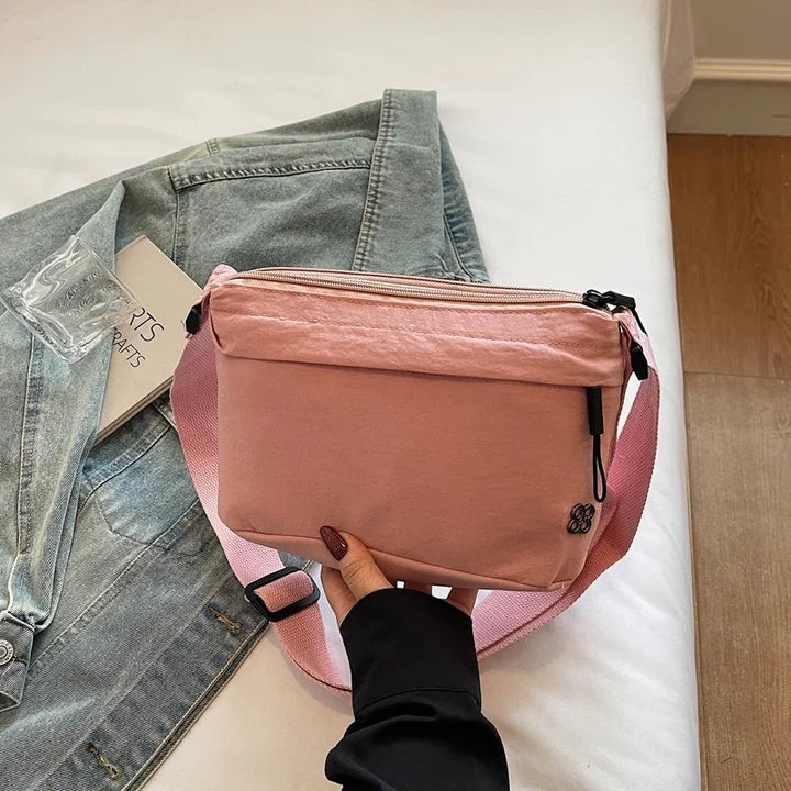 Pink crossbody bag held over denim jacket and book on white bed with wooden floor