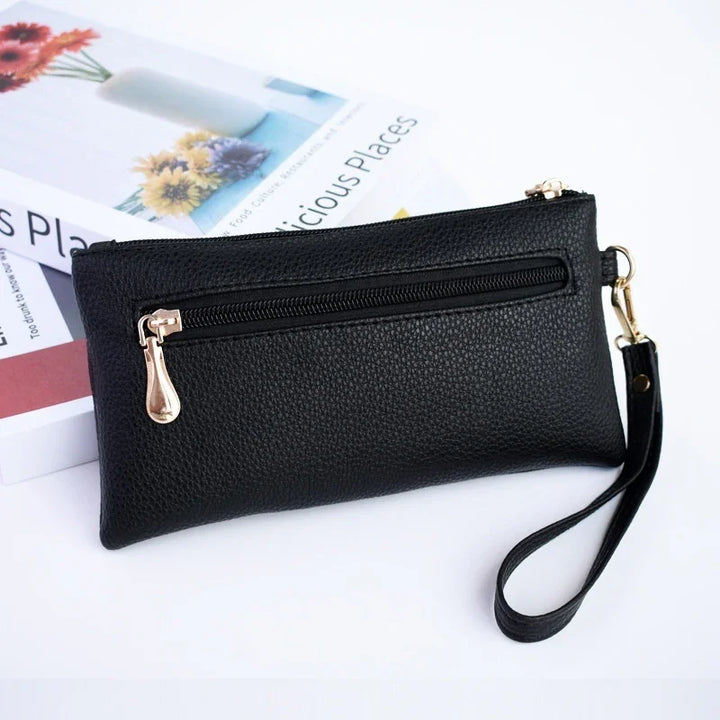 Black textured leather wristlet clutch with gold zipper and detachable strap, placed on books with floral covers