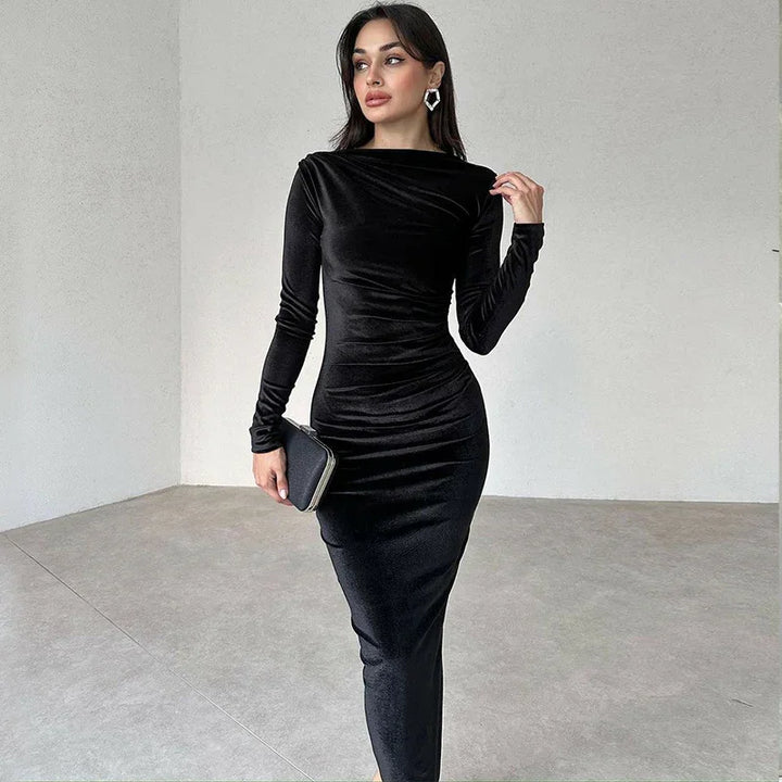 Woman wearing a fitted black long-sleeve velvet midi dress holding a black clutch in a minimalist room