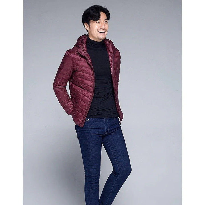 Smiling man posing in a maroon quilted jacket, black turtleneck, and blue jeans against gray background