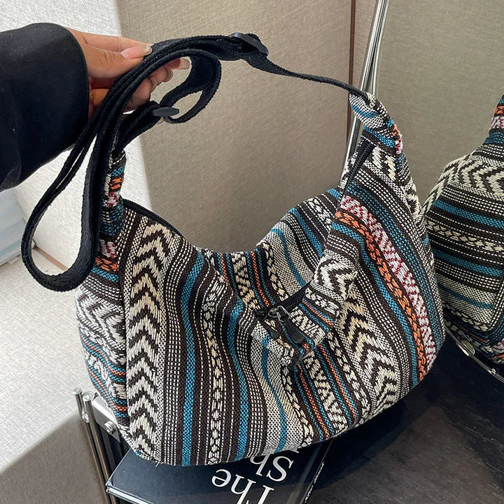 Hand holding a woven boho shoulder bag with black, blue, white, and orange striped pattern on a dark surface