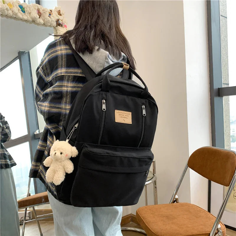 Person wearing black backpack with teddy bear keychain in casual indoor setting near window and brown chair
