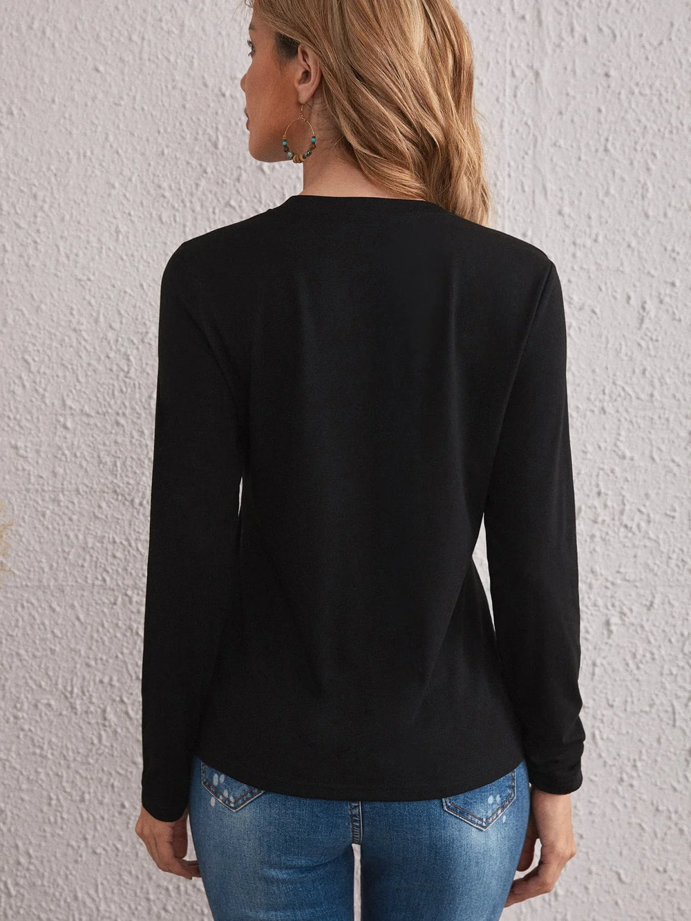 Woman wearing black long sleeve top and blue jeans standing against textured white wall