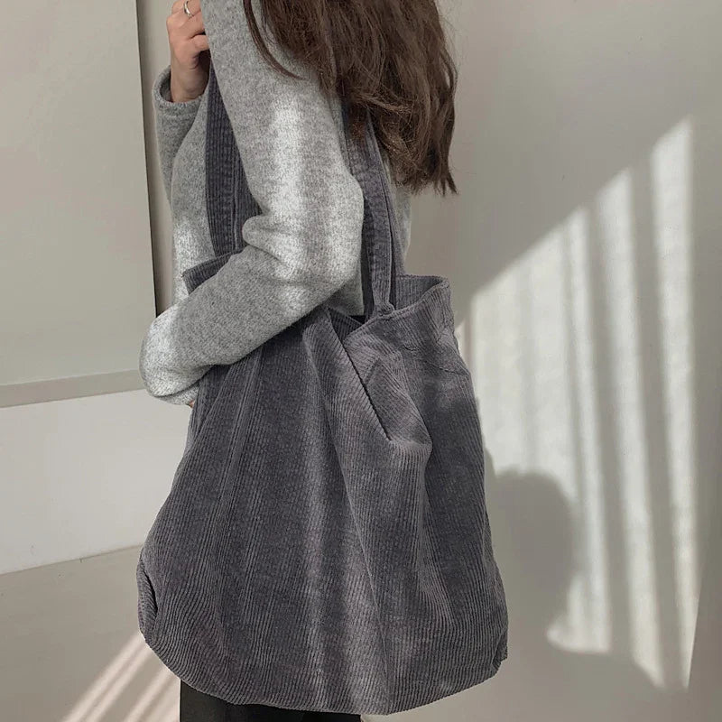 Woman wearing gray sweater carrying large dark gray corduroy tote bag in minimalist room