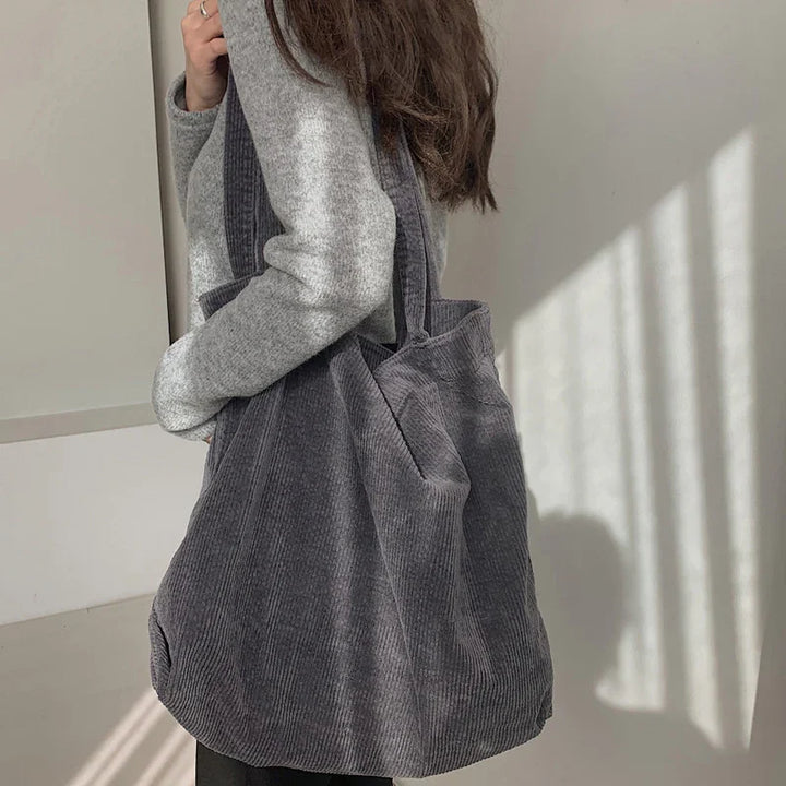 Woman wearing gray sweater carrying large dark gray corduroy tote bag in minimalist room
