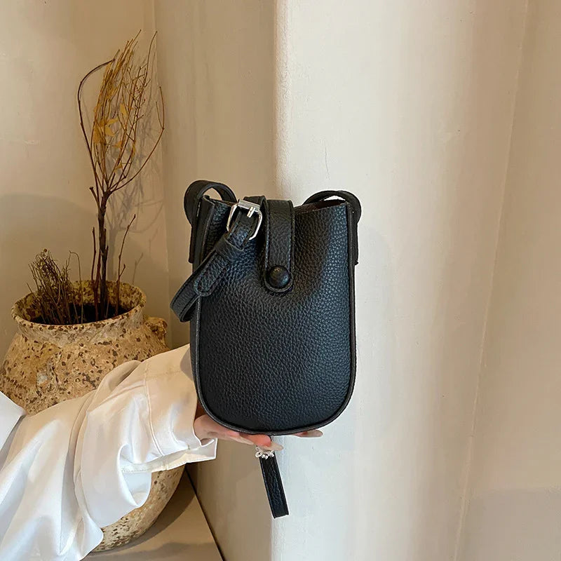 Hand holding small black leather crossbody bag with buckle strap in neutral indoor setting