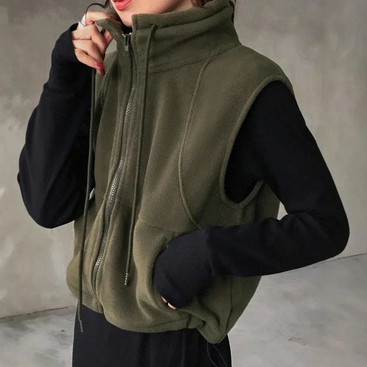 Person wearing olive green fleece zip-up vest over black long sleeves against gray wall