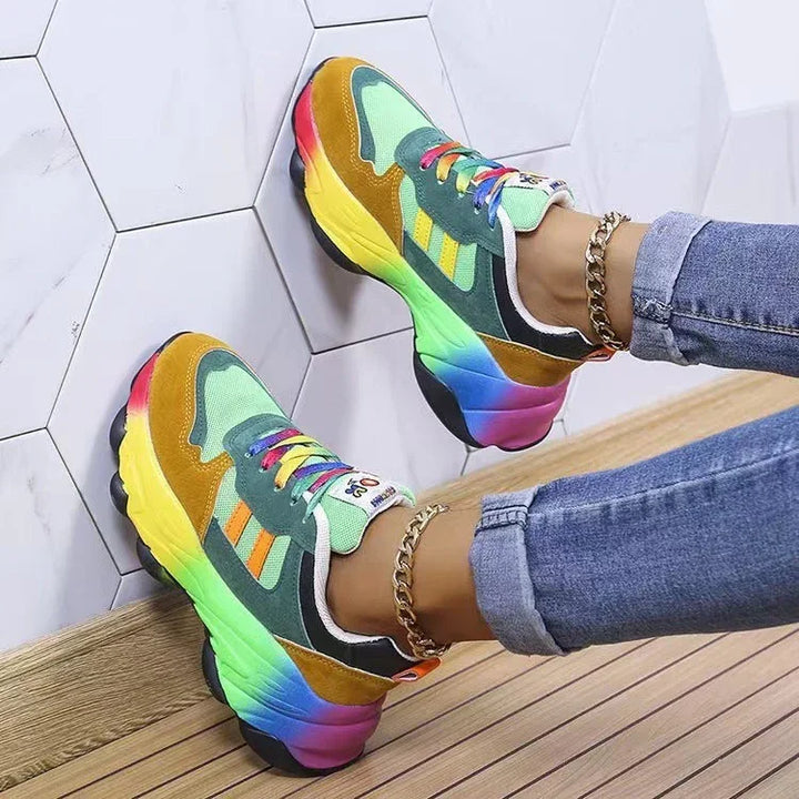 Colorful chunky sneakers with rainbow gradient soles worn with blue jeans and gold anklets on wooden floor and white hex tile wall