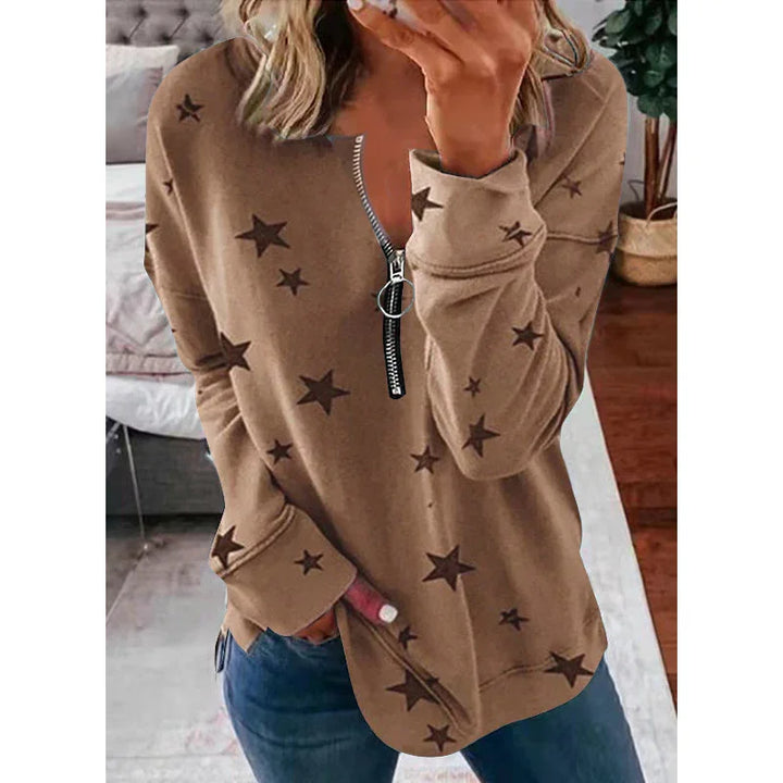 Woman wearing brown star print zip-up sweatshirt with blue jeans in cozy bedroom setting