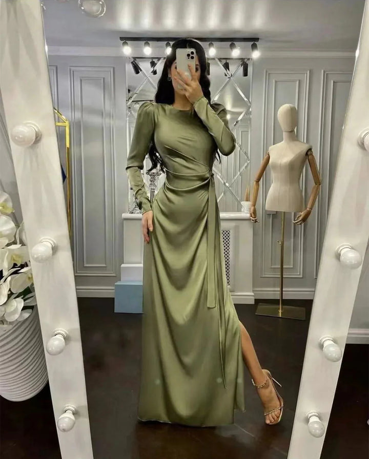 Woman in a long olive green satin dress with puff sleeves and side slit, posing in a modern dressing room