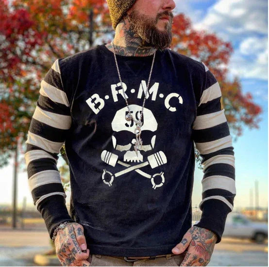 Bearded man with tattoos wearing a black B.R.M.C skull logo long-sleeve shirt outdoors with fall trees