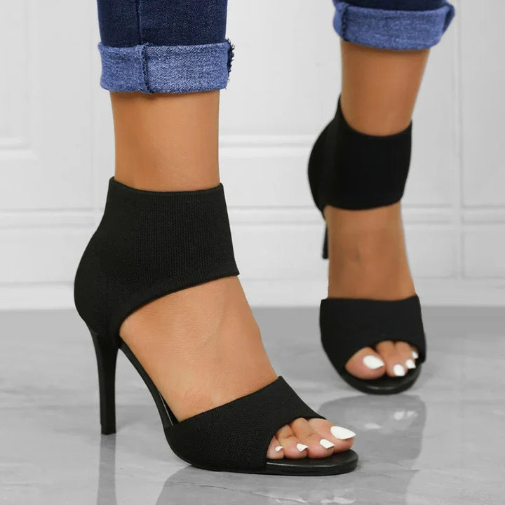 Women's black open-toe high heel ankle boots paired with rolled-up denim jeans on a marble floor