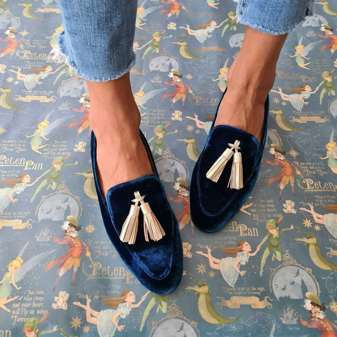 Feet wearing navy blue velvet loafers with beige tassels on Peter Pan-themed floor mat