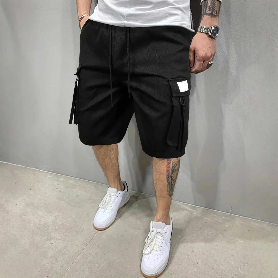 Man wearing black cargo shorts with side pockets and white sneakers, standing against gray wall