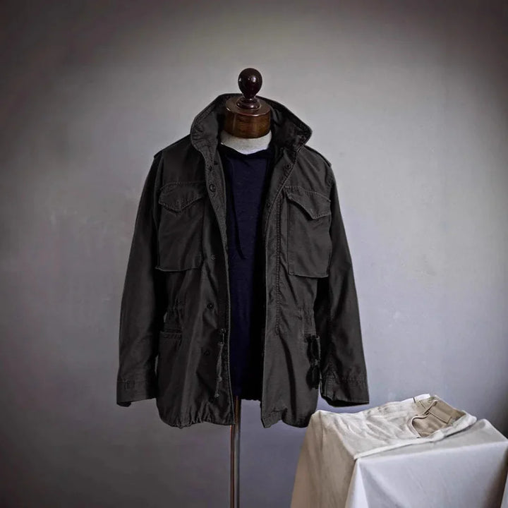 Mannequin wearing dark green military jacket over navy blue shirt with folded light beige pants on table