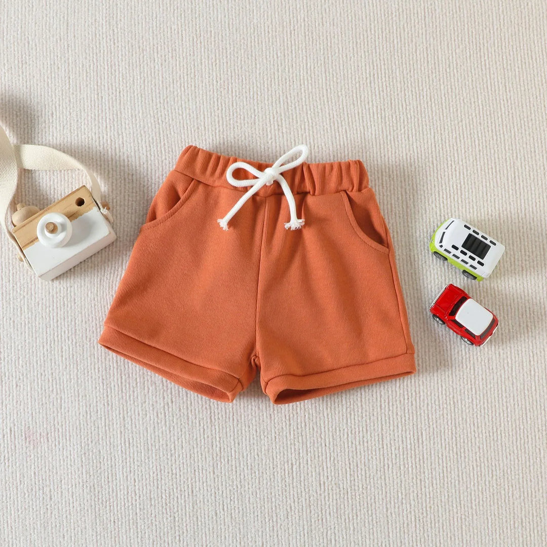 Rust-colored toddler shorts with white drawstring on textured beige background with wooden camera toy and small toy cars