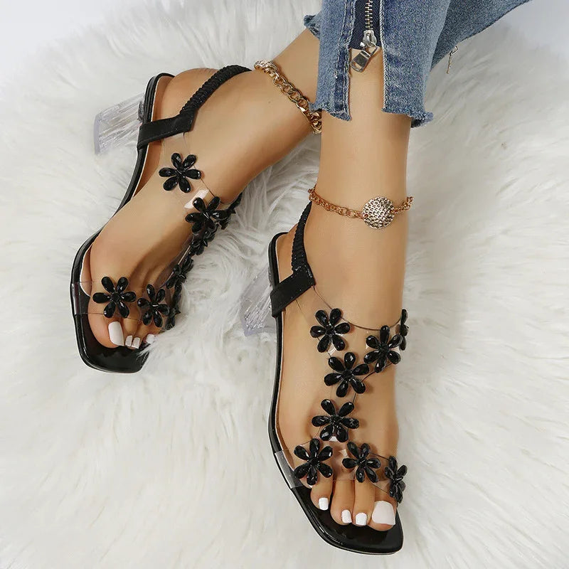 Women's black floral sandals with clear heels and ankle bracelets on feet in blue jeans