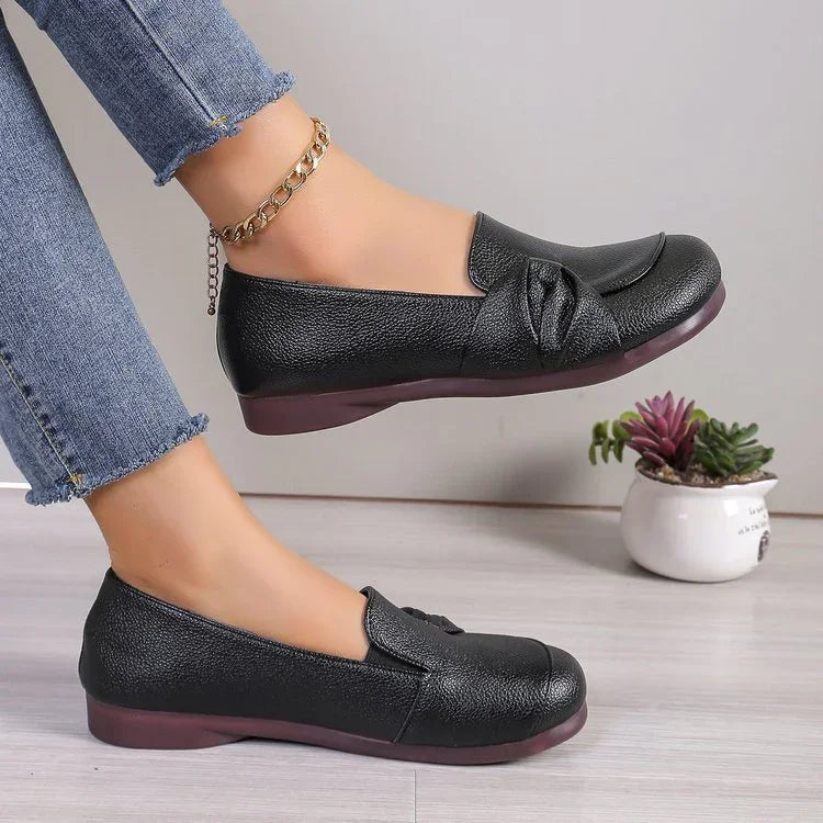 Black leather flat shoes with bow detail worn with blue jeans and gold anklet on light floor