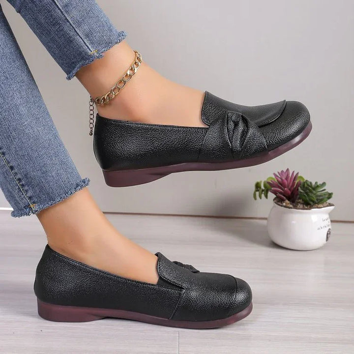 Black leather flat shoes with bow detail worn with blue jeans and gold anklet on light floor