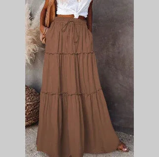 Woman wearing a brown tiered maxi skirt with drawstring waist and white knotted top against gray wall