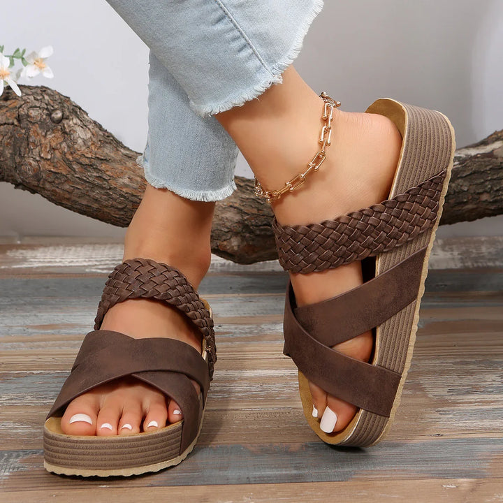 Close-up of feet wearing brown platform sandals with braided straps and gold anklet on wooden floor